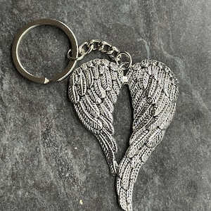 Puede incluir: Llavero plateado con alas de ángel. Las alas están detalladas con texturas de plumas y conectadas en el centro. Una pequeña cadena y un llavero redondo están unidos. El llavero es de metal.