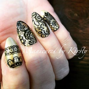 May include: Close-up of a hand with five nails painted in a gold color with black lace designs. The nails are long and pointed.