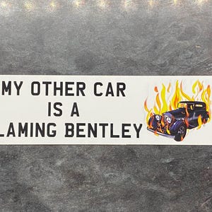 May include: White rectangular sticker with the text "MY OTHER CAR IS A FLAMING BENTLEY" in black. A graphic of a vintage black car engulfed in flames is on the right side. The sticker is on a gray surface.