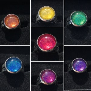 Può includere: Set di dieci anelli d'argento con glitter colorati incorporati al centro di ogni anello. Gli anelli sono disposti a griglia su uno sfondo nero. I colori del glitter sono rosso, arancione, giallo, verde, blu, viola, rosa e bianco.
