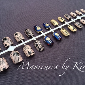 May include: A set of 18 false nails with an Egyptian theme. The nails are beige with black and gold designs. Some nails feature a blue and gold pharaoh's head design. The nails are displayed on a white plastic tray. "Manicures by Kirsty" is written in white cursive on a gray background.