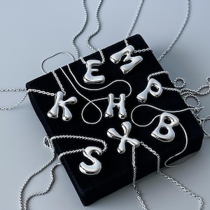 Puede incluir: Una colección de collares con iniciales plateadas expuestos sobre una superficie de terciopelo negro. Se ven las letras E, M, P, B, K, H, S y X, cada una suspendida de una delicada cadena. El fondo es gris.
