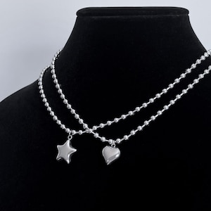 May include: Two silver chains with a star and heart pendant. The chains are layered on a black background.