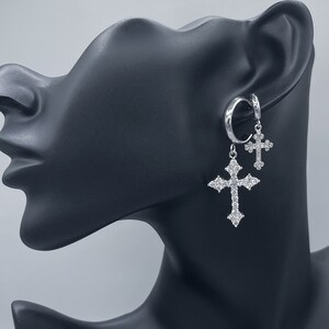 May include: A pair of silver hoop earrings with dangling cross charms. The crosses are adorned with clear cubic zirconia stones.