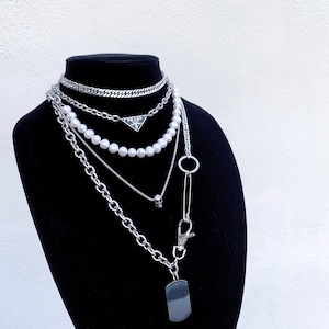 Puede incluir: Un conjunto de collares de plata en capas con una cadena de perlas, una cadena con un colgante de calavera, una cadena con un colgante de logotipo de Prada y una cadena con un colgante de placa.