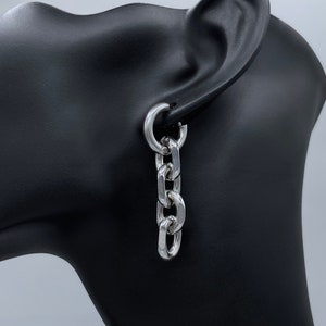 May include: Silver chain link earrings with a hoop closure. The earrings are hanging from a mannequin's ear.