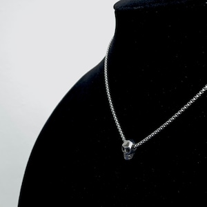 Puede incluir: Un collar de cadena de plata con un peque&ntilde;o colgante de calavera detallado. El collar se muestra en un soporte de terciopelo negro.