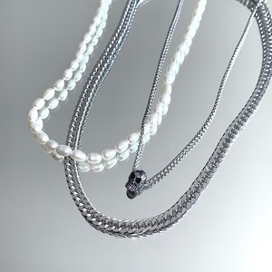 Puede incluir: Un collar de capas con una cadena de plata y una hebra de perlas. La cadena tiene un peque&ntilde;o colgante de calavera negra.