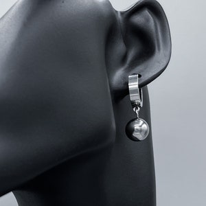 Puede incluir: Pendientes de aro de plata con una bola de plata colgante. La bola es lisa y brillante.