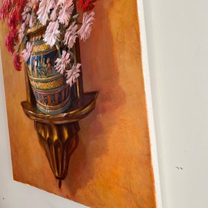 May include: A painting of a bouquet of pink and red flowers in a decorative vase on a wooden shelf. The vase is decorated with blue, yellow, and white geometric patterns. The background is a warm orange color. The painting is signed "Sidar Oliveira" in the lower right corner.