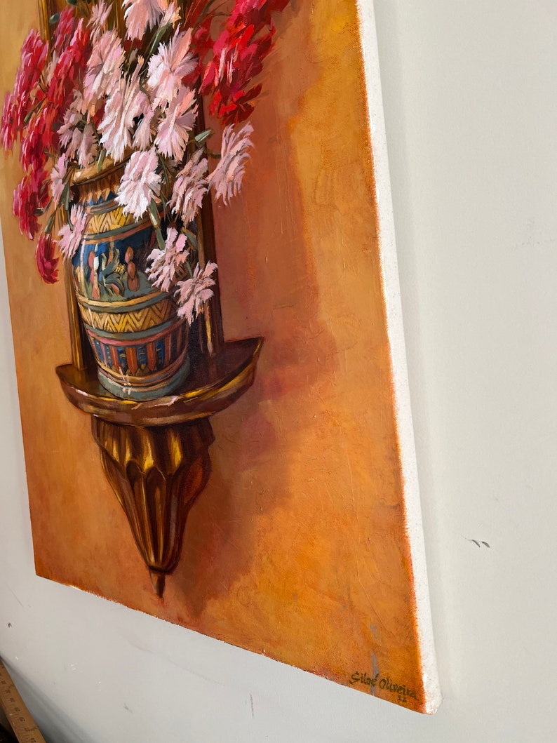 May include: A painting of a bouquet of pink and red flowers in a decorative vase on a wooden shelf. The vase is decorated with blue, yellow, and white geometric patterns. The background is a warm orange color. The painting is signed "Sidar Oliveira" in the lower right corner.