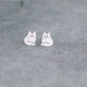 Puede incluir: Un par de pendientes de bot&oacute;n de plata con forma de gato. Los gatos miran hacia adelante con las colas enroscadas alrededor de sus cuerpos.