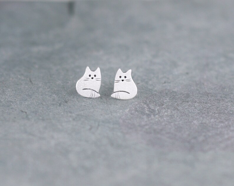 Puede incluir: Un par de pendientes de bot&oacute;n de plata con forma de gato. Los gatos miran hacia adelante con las colas enroscadas alrededor de sus cuerpos.