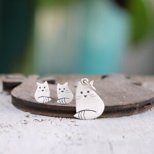 Puede incluir: Un conjunto de pendientes y un collar con colgante de gato de plata. El gato est&aacute; representado en un estilo simple y minimalista con un cuerpo redondeado y una cola peque&ntilde;a.