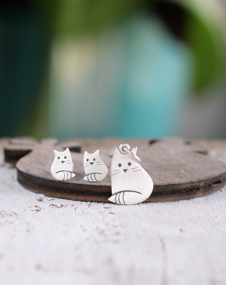 Puede incluir: Un conjunto de pendientes y un collar con colgante de gato de plata. El gato est&aacute; representado en un estilo simple y minimalista con un cuerpo redondeado y una cola peque&ntilde;a.