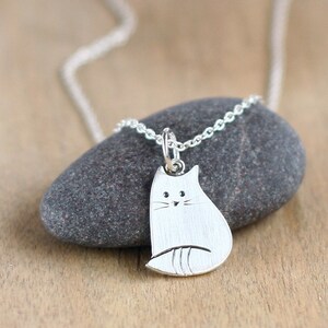 Puede incluir: Un collar de plata con un colgante de gato simple y minimalista. El gato est&aacute; representado en posici&oacute;n sentada con un cuerpo ligeramente redondeado y una peque&ntilde;a cara estilizada.