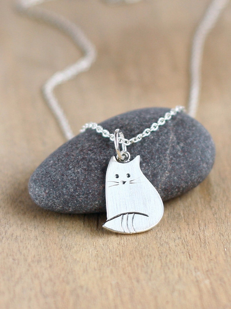 Puede incluir: Un collar de plata con un colgante de gato simple y minimalista. El gato est&aacute; representado en posici&oacute;n sentada con un cuerpo ligeramente redondeado y una peque&ntilde;a cara estilizada.