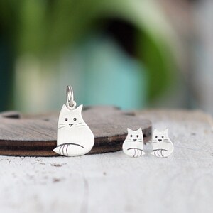 Puede incluir: Un conjunto de collar y pendientes de plata con un dise&ntilde;o de gato estilizado simple. El dije es m&aacute;s grande que los pendientes y cuelga de una cadena de plata.