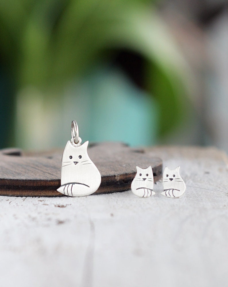 Puede incluir: Un conjunto de collar y pendientes de plata con un dise&ntilde;o de gato estilizado simple. El dije es m&aacute;s grande que los pendientes y cuelga de una cadena de plata.