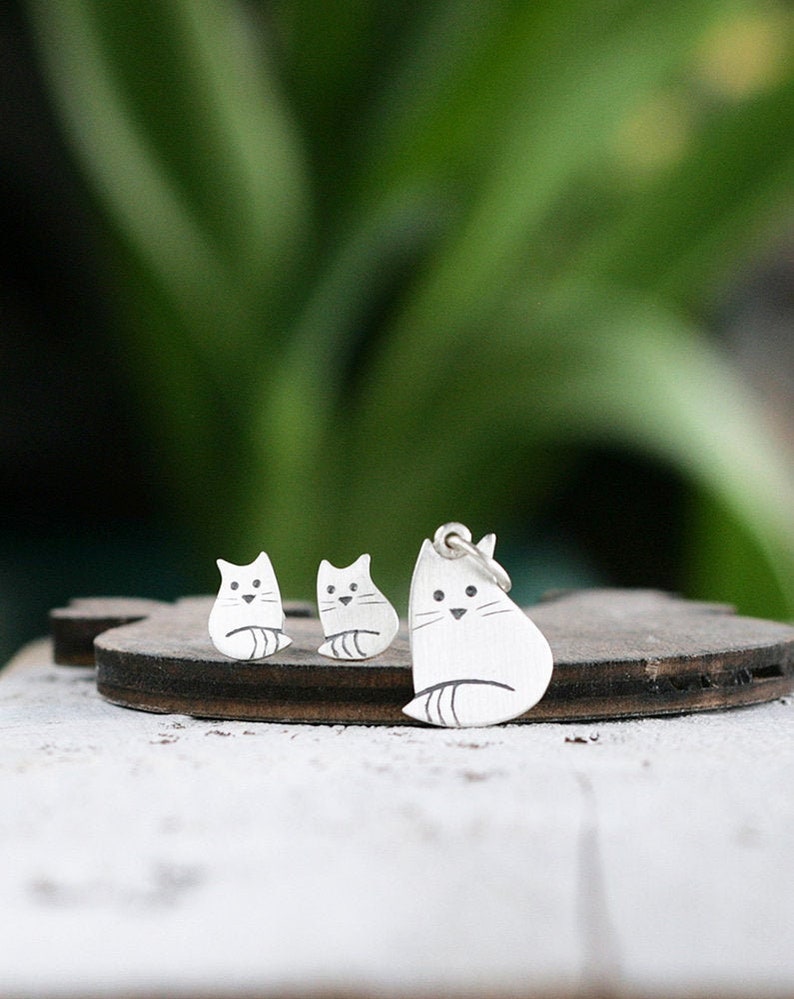 Puede incluir: Un conjunto de pendientes de plata con forma de gato y un collar con un colgante de plata con forma de gato. Los dijes de gato son simples y minimalistas, con una linda cara de estilo de dibujos animados.