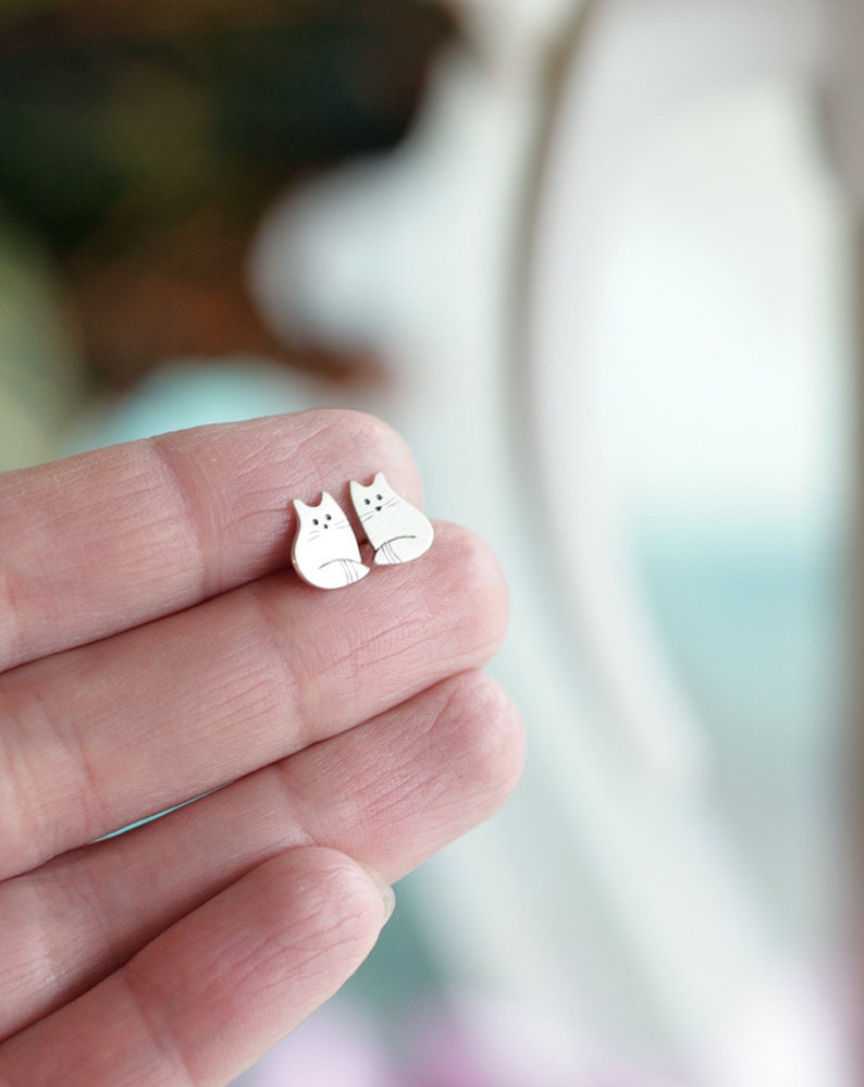 Puede incluir: Un par de pendientes de plata con forma de gato. Los gatos est&aacute;n mirando hacia adelante con sus colas enroscadas alrededor de sus patas.