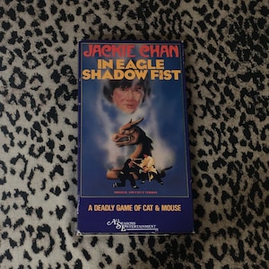 May include: A VHS tape featuring Jackie Chan in the movie "Eagle Shadow Fist". The cover features a blue background with a picture of Jackie Chan and a dragon. The text "A Deadly Game of Cat & Mouse" is printed on the bottom of the cover.