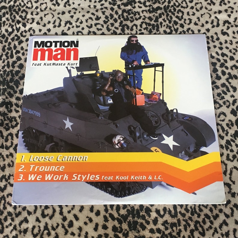 May include: A black and white image of a record cover featuring a tank with two men on top. The text on the cover reads "MOTION man feat Kut Masta Kurt" and lists the songs "Loose Cannon", "Trounce", and "We Work Styles feat Kool Keith & L.C."