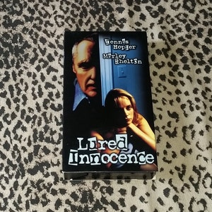 May include: VHS video cassette cover with a black and white image of a man and woman. The title of the movie is "Lured Innocence" and the actors are Dennis Hopper and Marley Shelton.