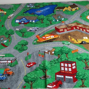 May include: A vibrant play mat depicting a town scene with roads, buildings, and vehicles. The mat includes a fire station, truck stop, and construction site. Cars, trucks, and a helicopter are also depicted on the green and grey surface.