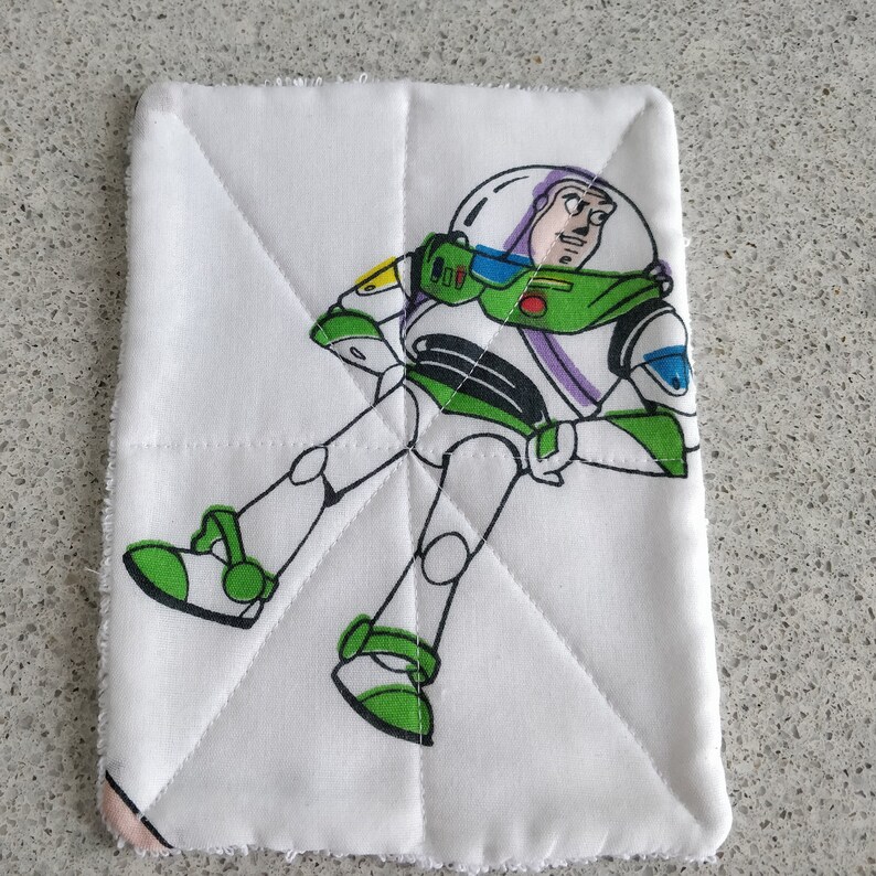 May include: White, quilted pot holder featuring a colorful illustration of Buzz Lightyear from Toy Story. The character is depicted in his signature green, white, and purple spacesuit. The pot holder has a stitched, diamond pattern.