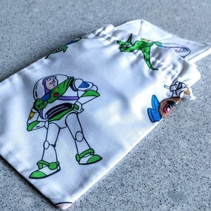 May include: White drawstring bag featuring Buzz Lightyear and other Toy Story characters in green, white, and blue. The bag is made of a silky material and is partially open, revealing the drawstring closure.