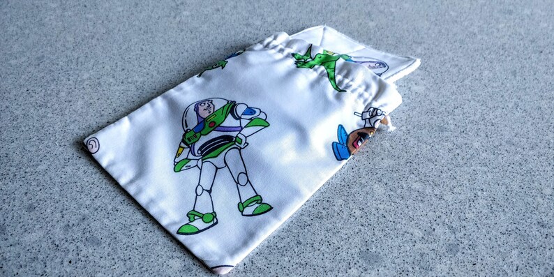 May include: White drawstring bag featuring Buzz Lightyear and other Toy Story characters in green, white, and blue. The bag is made of a silky material and is partially open, revealing the drawstring closure.