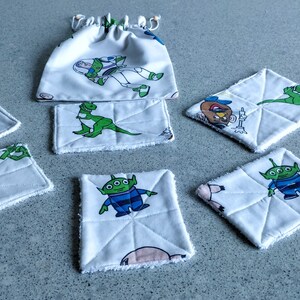 May include: A collection of white fabric items featuring characters from the animated film Toy Story. The set includes square and rectangular coasters, a drawstring bag, and a small rectangular item. Each piece has a different character design, including Buzz Lightyear and Mr. Potato Head.