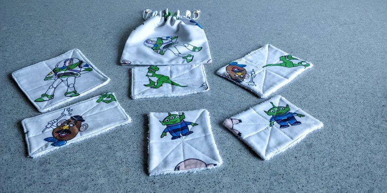 May include: A collection of white fabric items featuring characters from the animated film Toy Story. The set includes square and rectangular coasters, a drawstring bag, and a small rectangular item. Each piece has a different character design, including Buzz Lightyear and Mr. Potato Head.