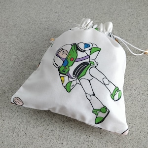 May include: White drawstring bag featuring a colorful illustration of Buzz Lightyear from Toy Story. The bag has a drawstring closure with wooden beads and is made of a soft, lightweight fabric. The design includes green, white, and purple accents.