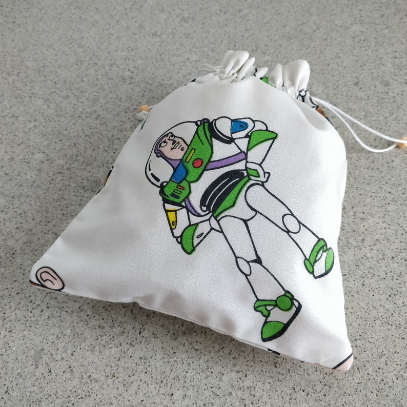 May include: White drawstring bag featuring a colorful illustration of Buzz Lightyear from Toy Story. The bag has a drawstring closure with wooden beads and is made of a soft, lightweight fabric. The design includes green, white, and purple accents.