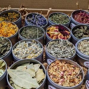 May include: A selection of dried herbs and flowers presented in small, round metal tins within a cardboard box. The tins contain various botanicals like lavender and chamomile. The arrangement displays a range of colours and textures, ideal for culinary or medicinal use.