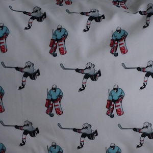 Peut inclure: Tissu blanc avec un motif répété de joueurs de hockey en uniforme bleu et rouge. Les joueurs tiennent des bâtons de hockey et portent des jambières de gardien de but. L'arrière-plan est blanc.