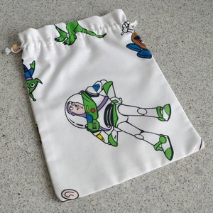 May include: White drawstring bag featuring cartoon characters from the Toy Story movie franchise. The bag has a repeating pattern of Buzz Lightyear, Mr. Potato Head, the Little Green Men, and Rex the dinosaur. The bag is made of a soft fabric and has a wooden bead on the drawstring.