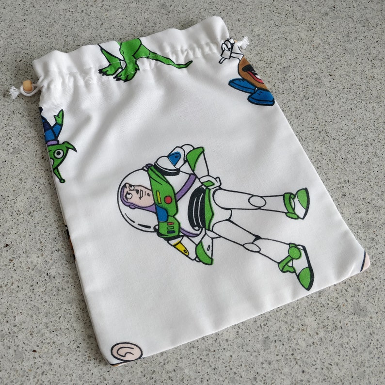 May include: White drawstring bag featuring cartoon characters from the Toy Story movie franchise. The bag has a repeating pattern of Buzz Lightyear, Mr. Potato Head, the Little Green Men, and Rex the dinosaur. The bag is made of a soft fabric and has a wooden bead on the drawstring.