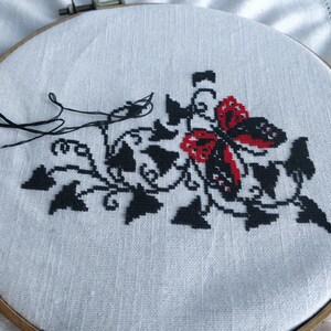 May include: Cross-stitch embroidery on white fabric, featuring a black and red butterfly design with black foliage. The embroidery is stretched within a wooden hoop, with black thread and needle visible.