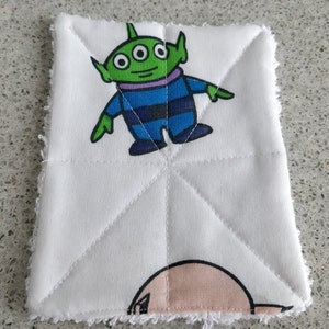 May include: White, square-shaped cloth featuring a cartoon alien character with three eyes, green skin, and a blue and black outfit. The cloth has a quilted pattern and a white, fringed edge.