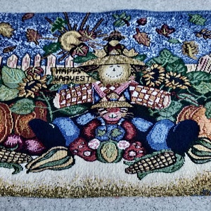May include: A colourful tapestry depicting a scarecrow sitting in a garden with pumpkins, corn, and sunflowers. The scarecrow is wearing a blue shirt, red trousers, and a straw hat. The tapestry features a white picket fence and a blue sky with clouds. The text "Happy Harvest" is written on a banner above the scarecrow.
