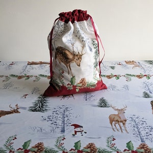 May include: A drawstring gift bag featuring a white background with a printed deer, pine branches, and pinecones. The top and bottom sections of the bag are a solid deep red fabric, and the bag is cinched at the top with matching red ribbons.