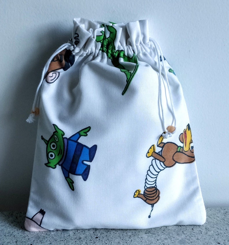 May include: White drawstring bag featuring colorful cartoon character illustrations. The bag has a cinched top with a white drawstring and wooden bead accents. The bag is made of a soft fabric.