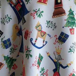 May include: A white fabric featuring a repeating Christmas-themed pattern of nutcrackers, blonde figures in blue dresses, rocking horses, green Christmas trees, wrapped gifts, drums, and sprigs of mistletoe in red, green, blue, and gold colors.