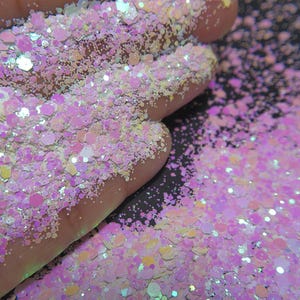 May include: Close-up of iridescent glitter in shades of pink, white, and yellow. The glitter is in various shapes and sizes, with a sparkling effect. The glitter is scattered on a dark surface.
