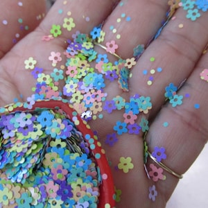 May include: A hand holding a container of flower-shaped confetti in pastel shades of blue, purple, pink, and yellow. Some confetti is scattered on the hand and fingers. The container has a red rim with white polka dots.
