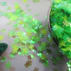 May include: Green iridescent glitter in the shape of four-leaf clovers. The glitter is loose and scattered on a white surface. A small amount of glitter is in a silver measuring spoon.