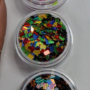 May include: Three clear plastic pots filled with colourful, iridescent glitter. The glitter pieces are rectangular and reflect various colours, including green, purple, orange, and blue. The pots are arranged vertically against a white background.